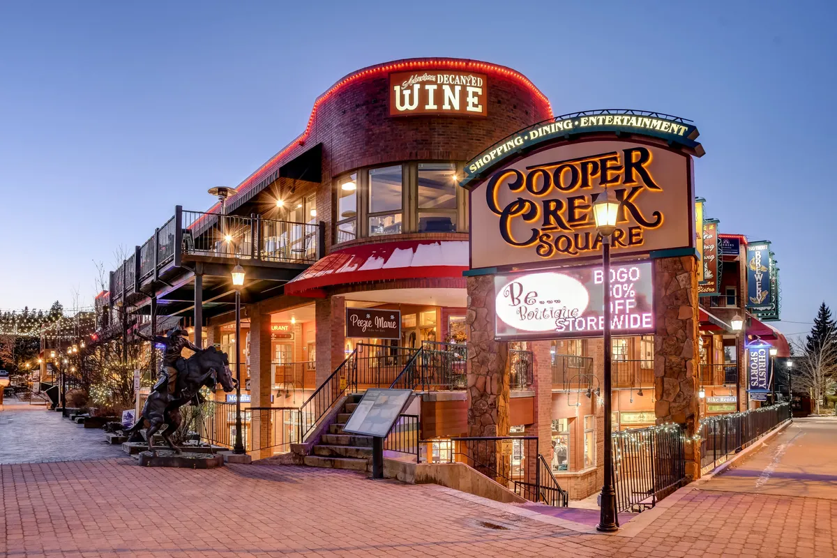 A festive streetscape featuring the Cooper Creek Square sign, a wine shop, and decorative lighting. The area is bustling with activity, inviting guests to explore local dining, shopping, and entertainment in the heart of downtown Winter Park.
