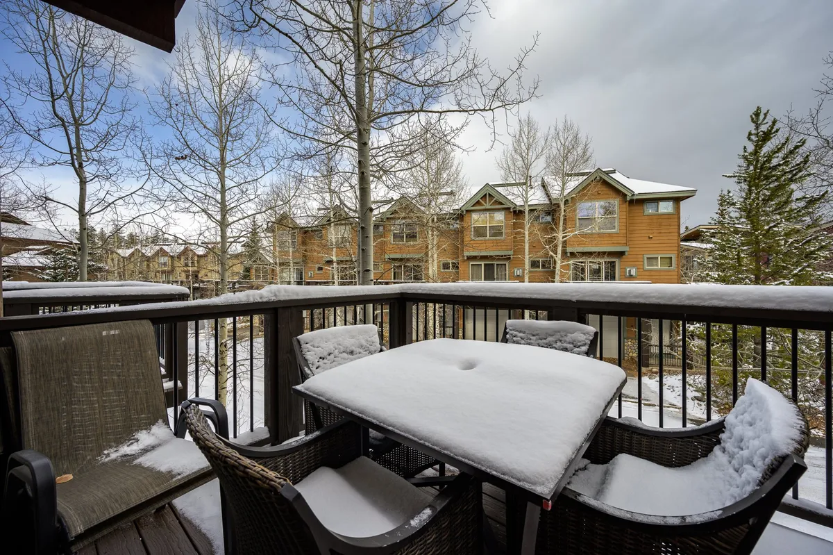 A snow-covered outdoor deck with comfortable furniture, a table dusted with fresh snow, and towering evergreen trees in the background. Neighboring buildings are visible, giving a sense of community and access to outdoor shared spaces.