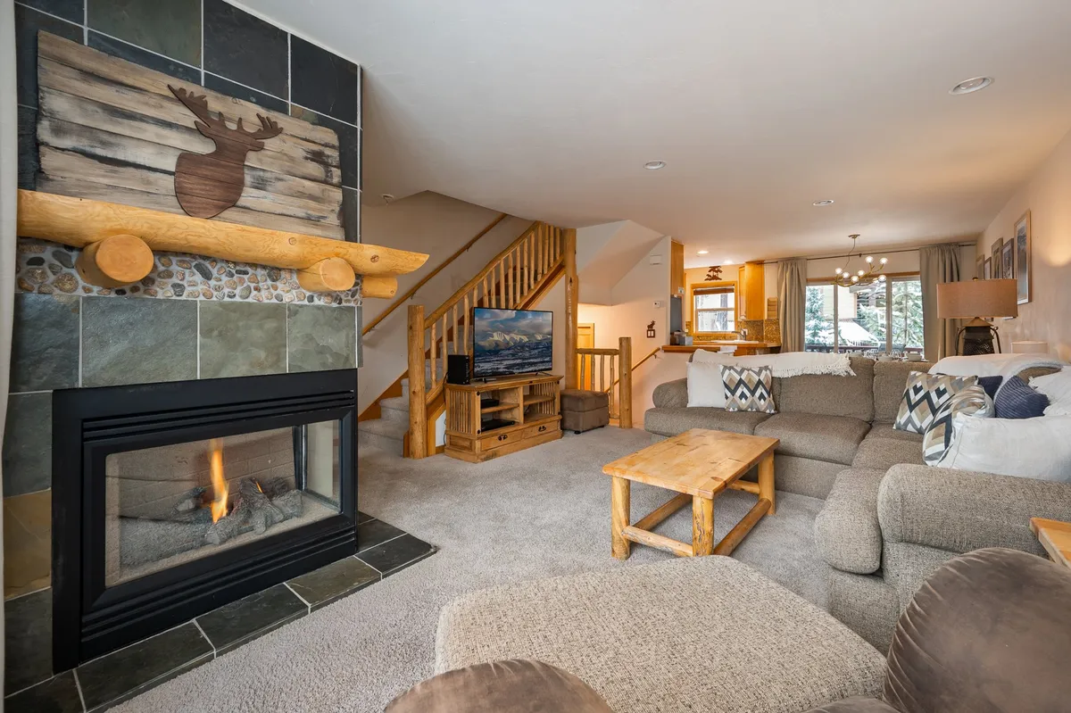 The image showcases a warm, inviting living space featuring a crackling fireplace, plush sofa, rustic wooden table, and modern staircase. The cozy arrangement is perfect for gathering, relaxing by the fire, or enjoying a movie night, all set against the backdrop of a stylish mountain townhome.