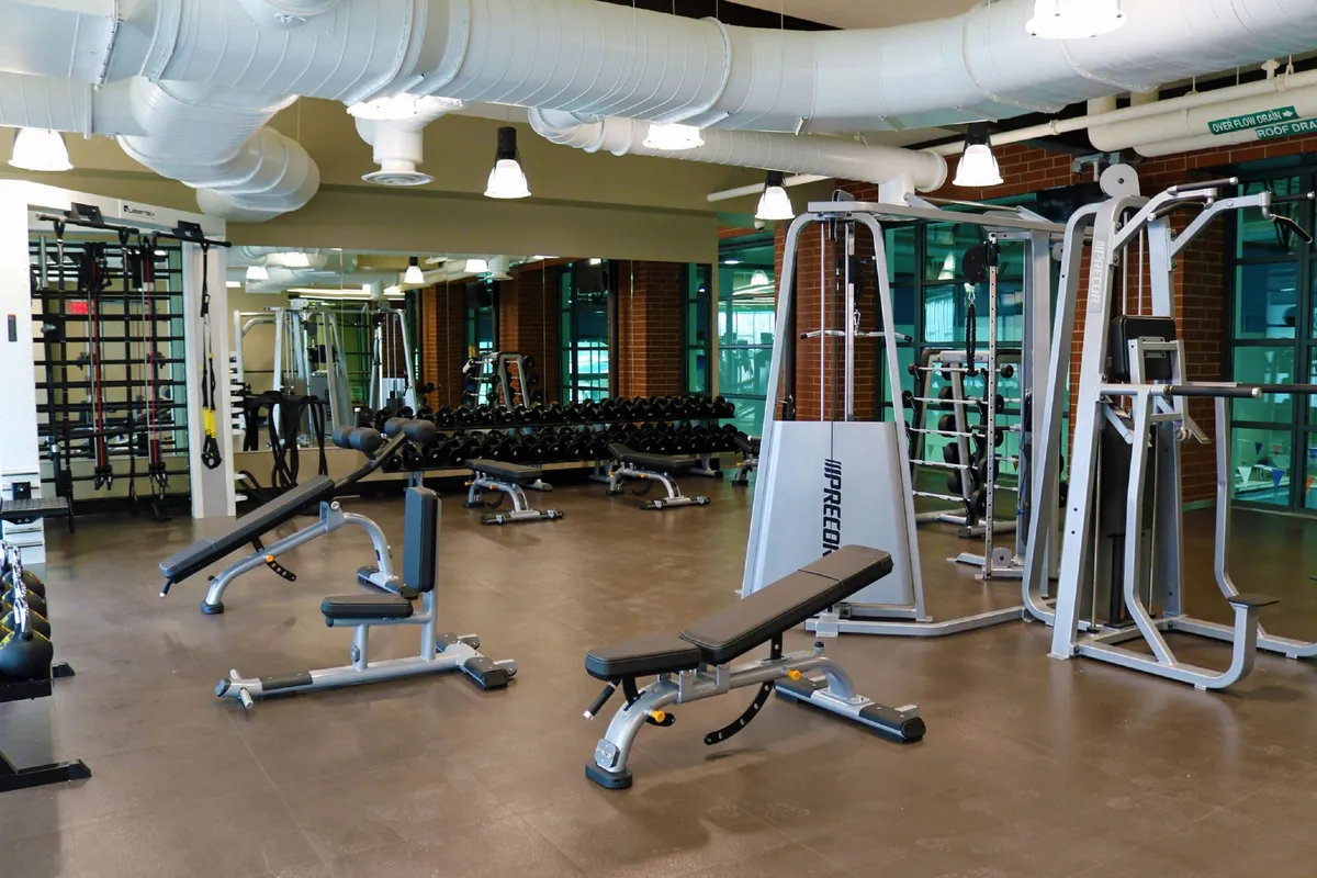 A dynamic fitness center scene featuring weight benches, dumbbell racks, and various exercise machines. The space is well-lit and modern, indicating access to upscale fitness and recreation facilities.