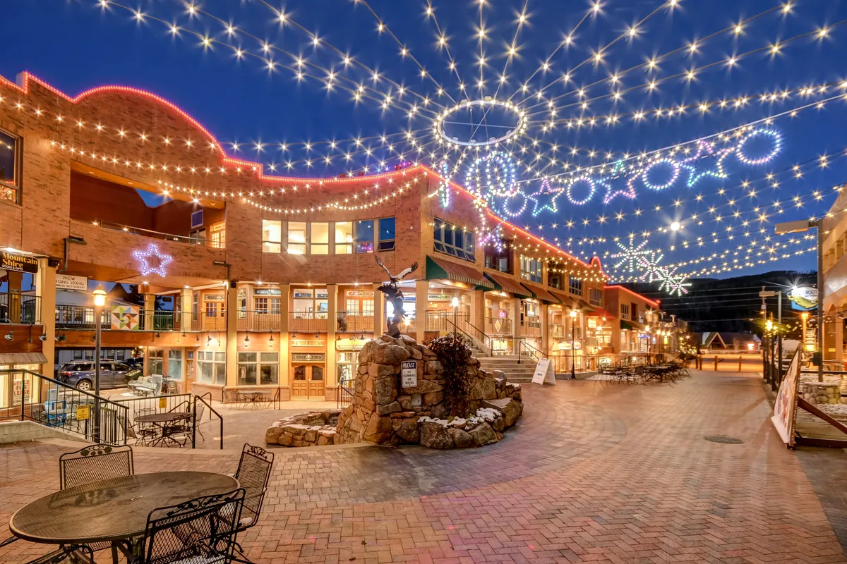 A lively exterior scene with string lights overhead, decorative stars, a stone fountain, and bustling cafes and shops. The festive mood suggests socializing, dining, and enjoying Winter Park's vibrant downtown energy just moments away.