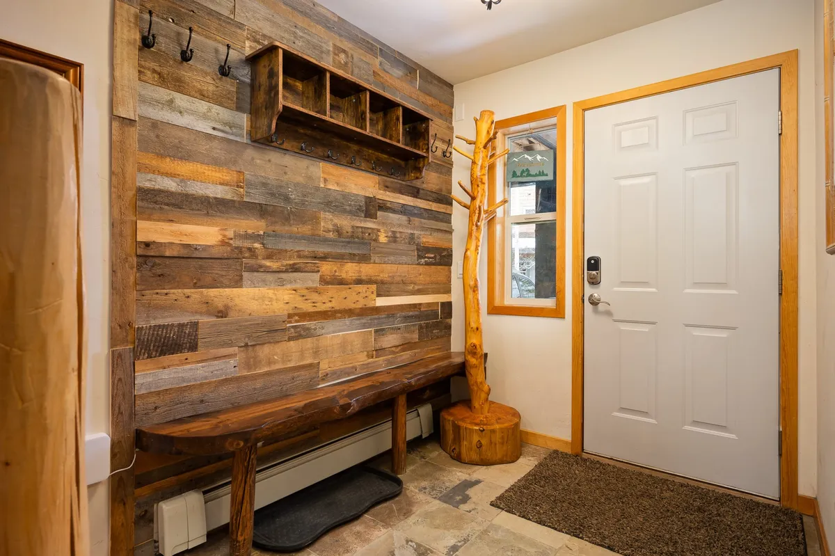 A cozy entryway featuring a wooden bench, rustic wall, a natural wood tree sculpture, and hooks for coats. The space is designed for welcoming guests, storing shoes, and hanging outerwear, providing a warm and practical first impression.