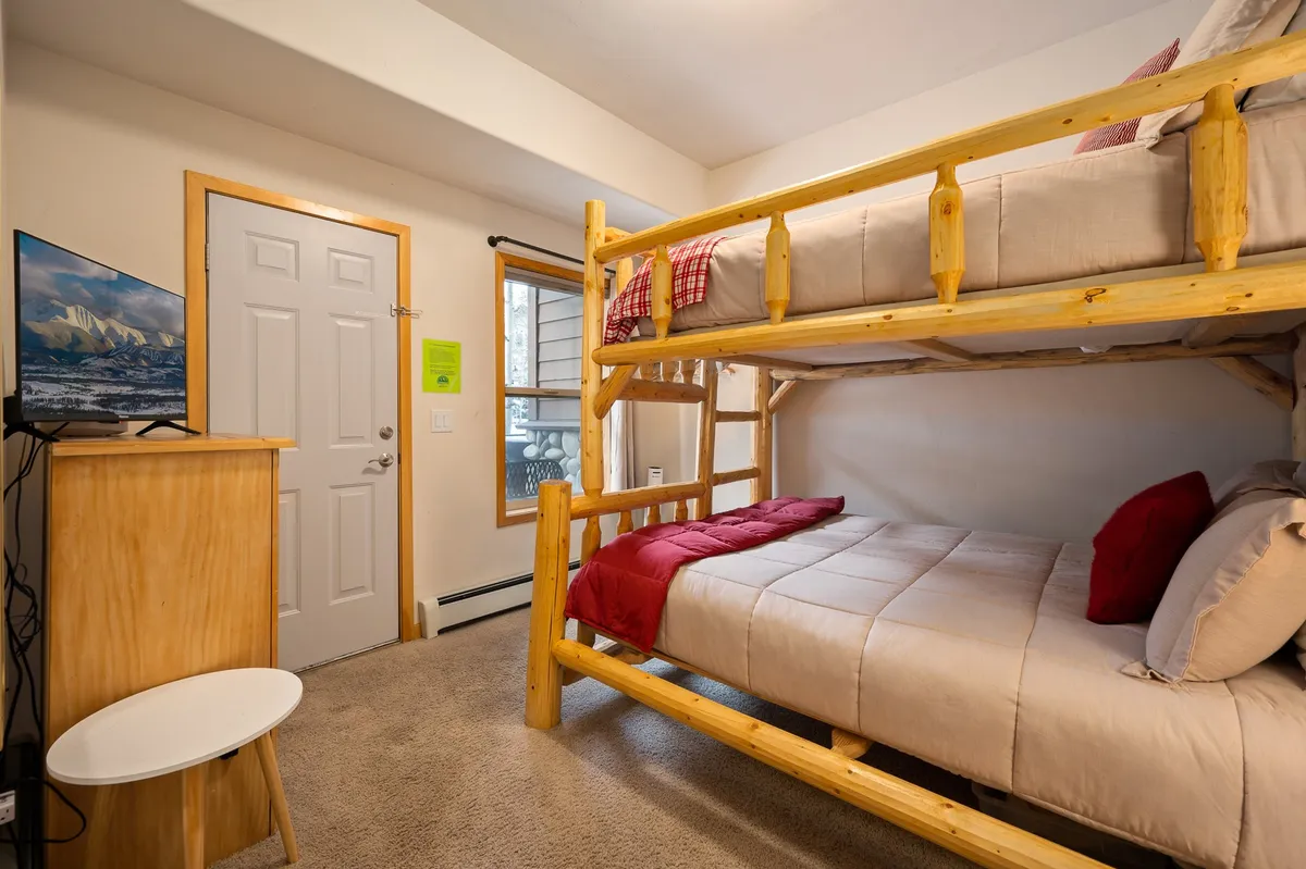 A cozy bunk room with full-over-full bunk beds, a television mounted on the wall, and a window with a scenic view. The room is designed for both comfort and fun, suitable for children, teens, or friends.