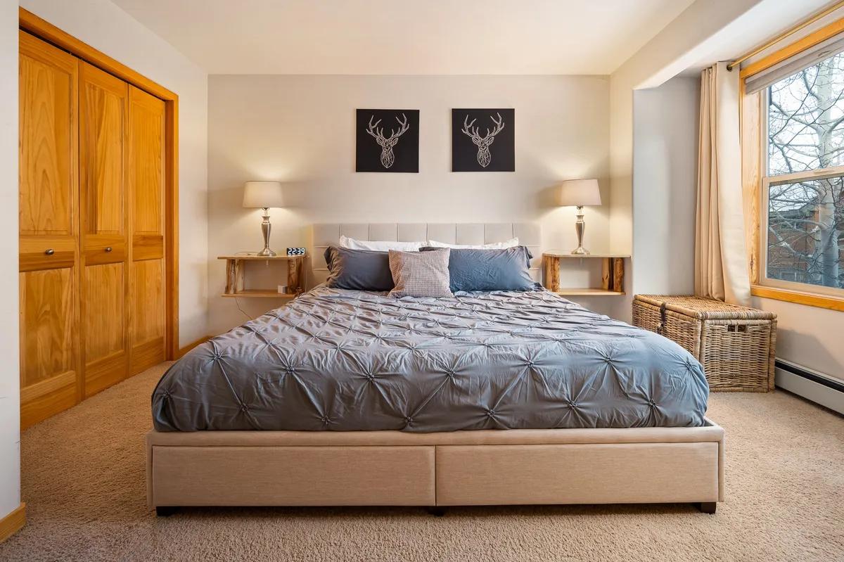 This image features a serene bedroom with a neatly made bed, soft lighting, a window allowing in natural light, and tasteful wall art. The overall ambiance is calm and inviting, ideal for relaxation and restful sleep.
