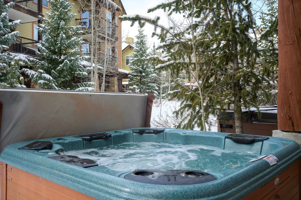 An inviting outdoor hot tub nestled on a private patio, surrounded by snow-covered trees and set against a beautiful mountain backdrop. The facade of the building is visible, and the tranquil scene suggests a soothing place to relax after a day of adventure.