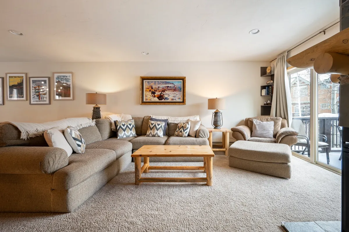 A large, inviting living room bathed in natural light from floor-to-ceiling windows. The space features a plush sectional sofa, a wooden coffee table, mountain-inspired artwork, and expansive windows that frame breathtaking views. The setup encourages relaxation, family movie nights, and socializing by bringing everyone together in comfort.